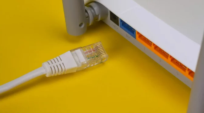 Roteador branco semelhante a um aparelho Simet Box, com cabo ao lado, em um fundo amarelo.